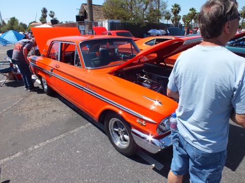 Orange Ford Fairlane hardtop at outdoor car show with onlooker