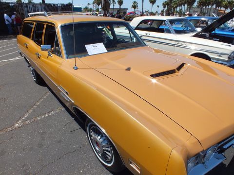 Gold station wagon with hood scoop and roof rack at a sunny car show