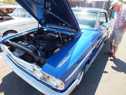 Blue classic car with open hood showing V8 engine, man in patterned shirt nearby