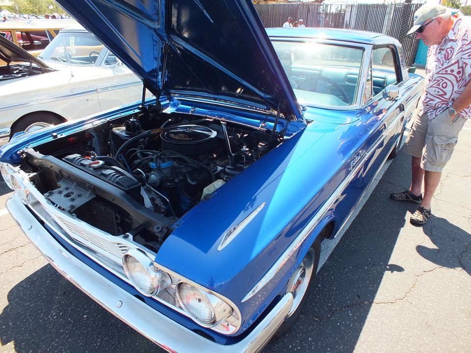 Blue classic car with open hood showing V8 engine, man in patterned shirt nearby
