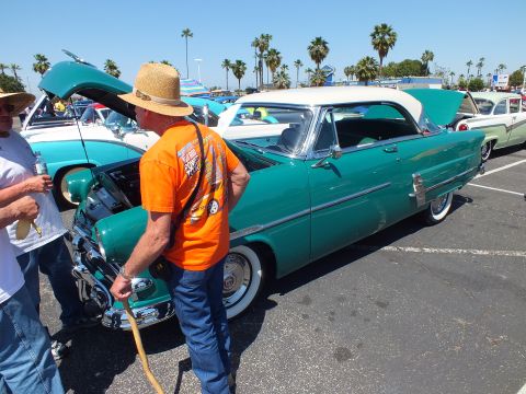 Man in orange shirt and straw hat beside teal 1950s hardtop at car show