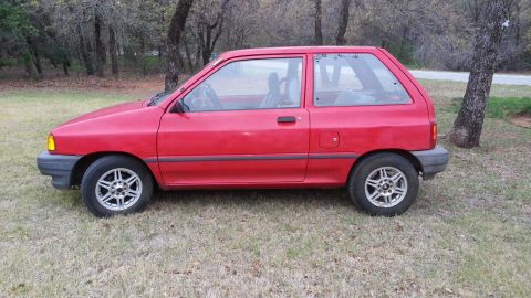 1991 Ford Festiva - A red hatchback is parked on a grassy area with trees in the background.