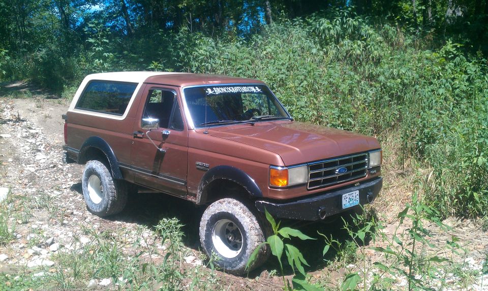 Brown 1987 Ford Bronco with a white top parked on a dirt road in a wooded area