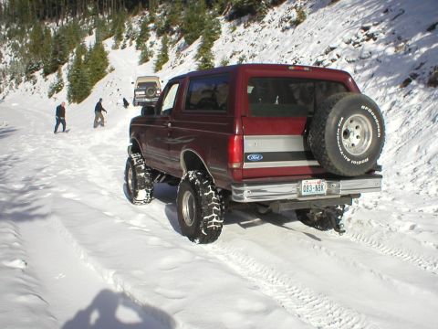 1978 Ford Bronco - A group of people are snowboarding down a snowy hill while a lifted Ford Bronco drives on the road below.