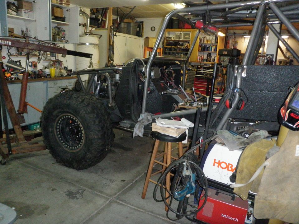 Off-road buggy frame with large tire, roll cage, and engine components in a garage