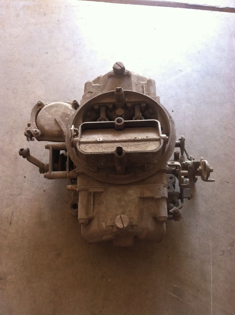Dusty carburetor with visible linkages and screws, resting on a concrete surface