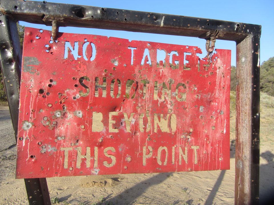 Red metal sign with bullet holes reading "NO TARGET SHOOTING BEYOND THIS POINT"