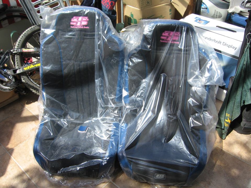Two racing seats in plastic wrap, one with pink "SS Twisted Stitch Seats" logo