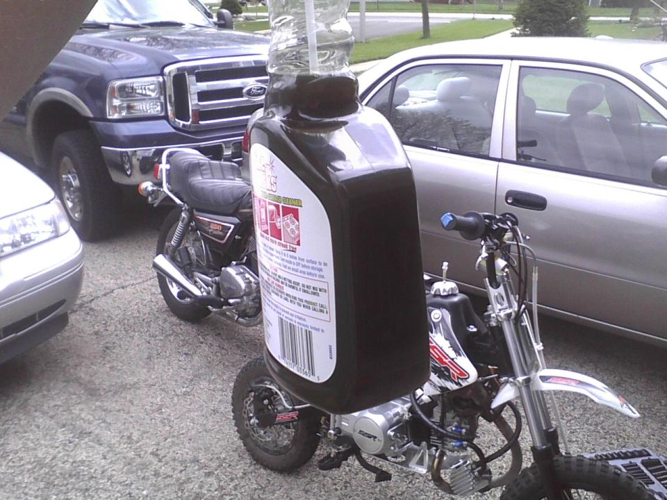 Motor oil bottle mounted on a dirt bike, with a classic motorcycle and Ford truck in the background