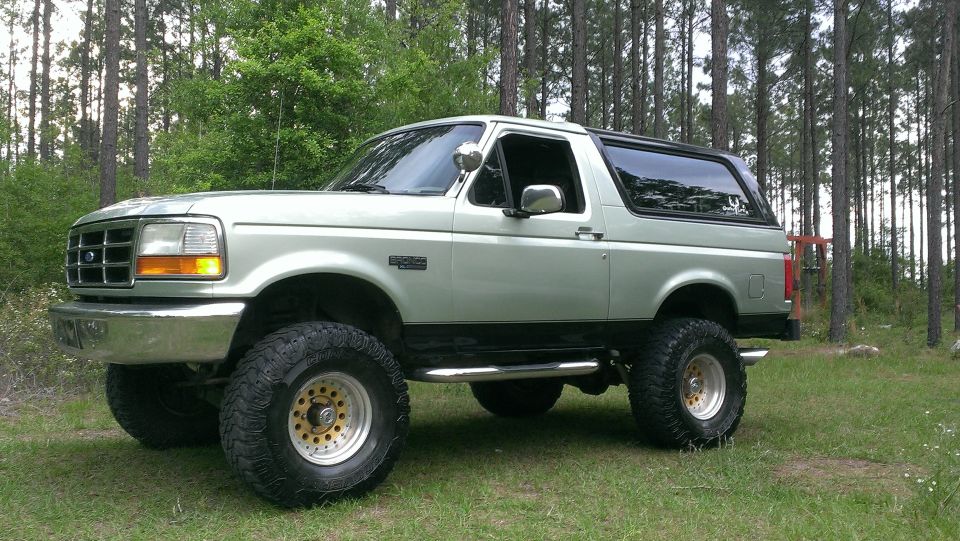 Silver 1995 Ford Bronco with lifted suspension and off-road tires parked in a wooded area