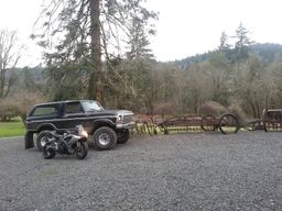 1979 Ford Bronco - A black Ford Bronco and a motorcycle sit in a gravel lot with trees and hills in the background.