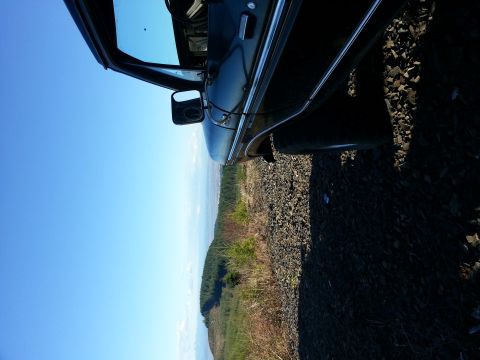 1979 Ford Bronco - Side view of a dark SUV door and mirror, parked on gravel with a wooded hillside and blue sky.