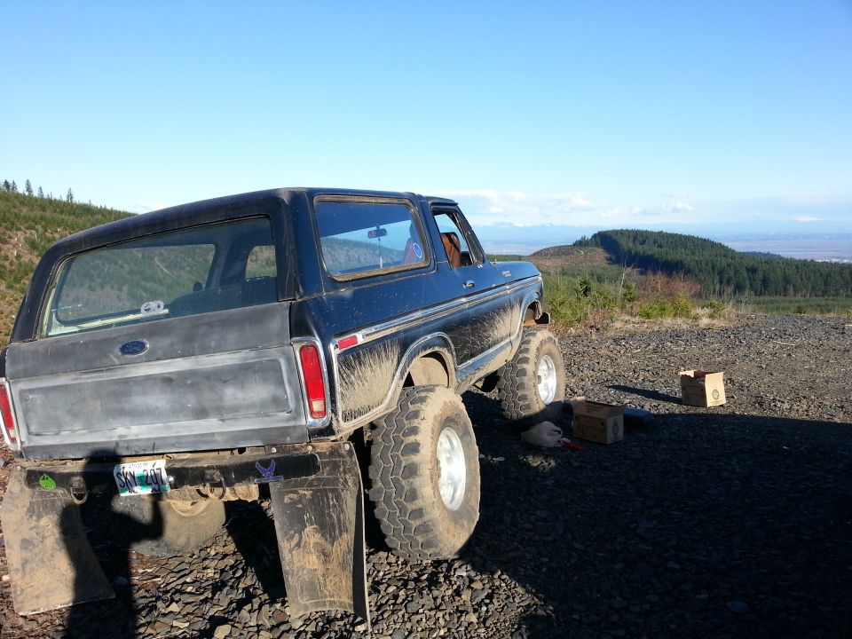 Dark blue 1978 Ford Bronco with lifted suspension and large tires, parked on a gravelly surface with a wooded hillside in the background