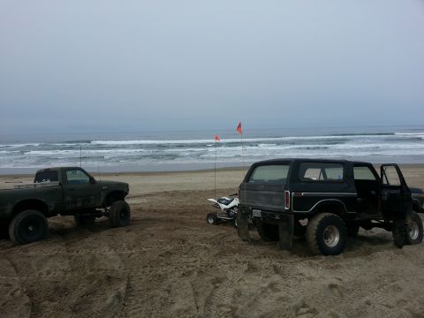 1979 Ford Bronco - A black 1979 Ford Bronco sits on a sandy beach with its door open, next to a quad bike and a pickup truck.