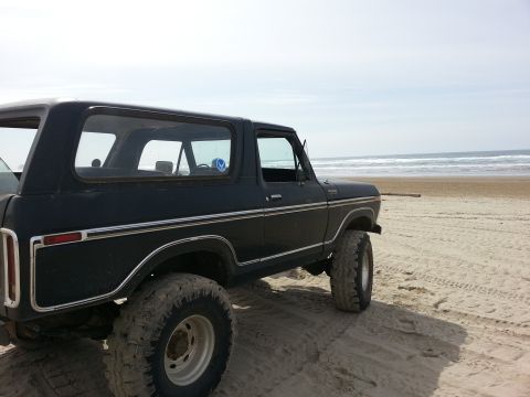 1979 Ford Bronco - SUV parked on a beach, showing its side profile and large off-road tires against the ocean backdrop.