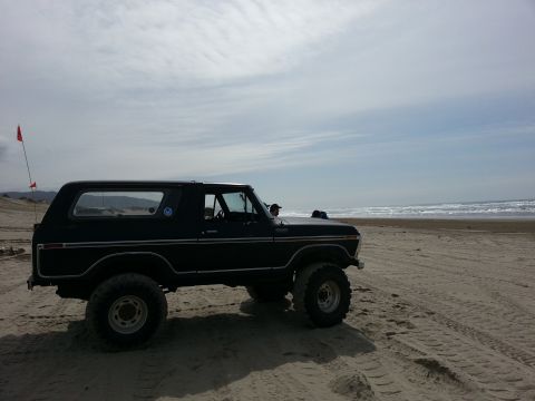 1979 Ford Bronco - A classic Ford Bronco sits on the sand with the ocean in the background under a cloudy sky.