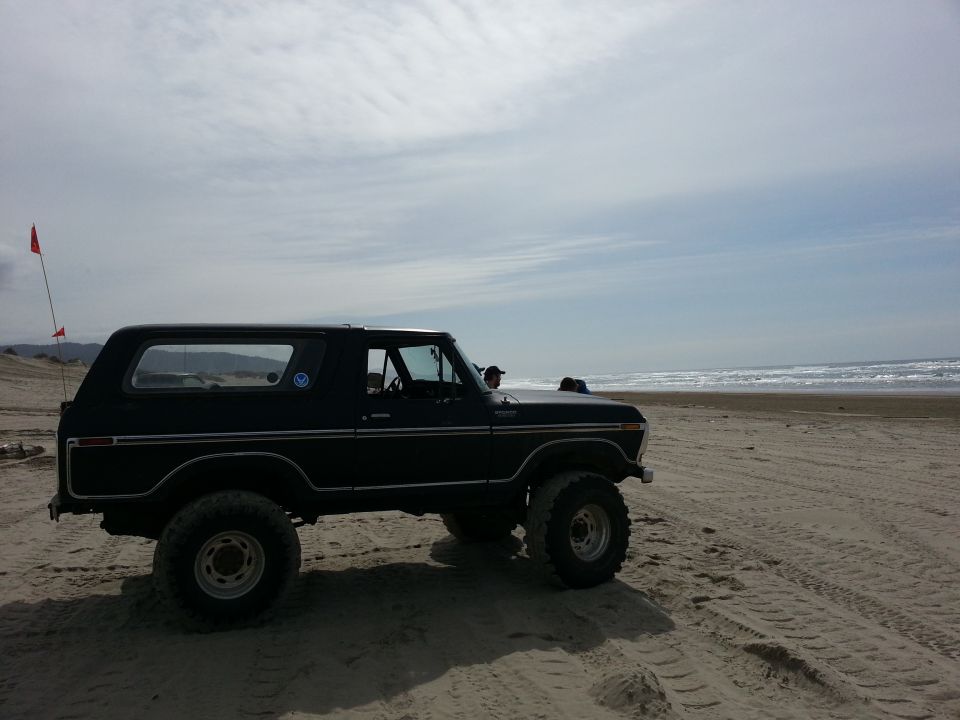 Black Ford Bronco parked on sand near ocean waves. Red flag on pole in background