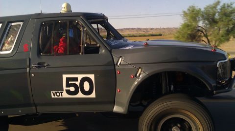 1996 Ford Bronco - Gray off-road prerunner truck shown in 3/4 front profile, featuring a caged cab with a red-suited occupant, roof-mounted white beacon, tubu…