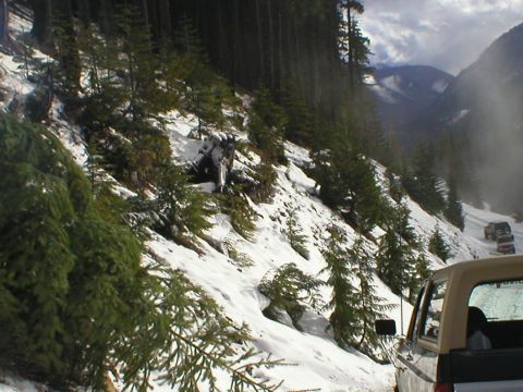 1978 Ford Bronco - Several SUVs drive along a snow-covered mountain road with dense pine trees on the slope.