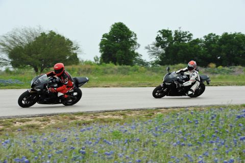 2001 Kawasaki KZ1000-A1 - Two sport motorcycles race on a track bordered by bluebonnets, with one rider in red and the other in white racing suits.