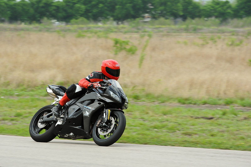 Motorcyclist in red and black gear on a black sportbike, leaning into a turn on a paved road with dry grass and trees in the background
