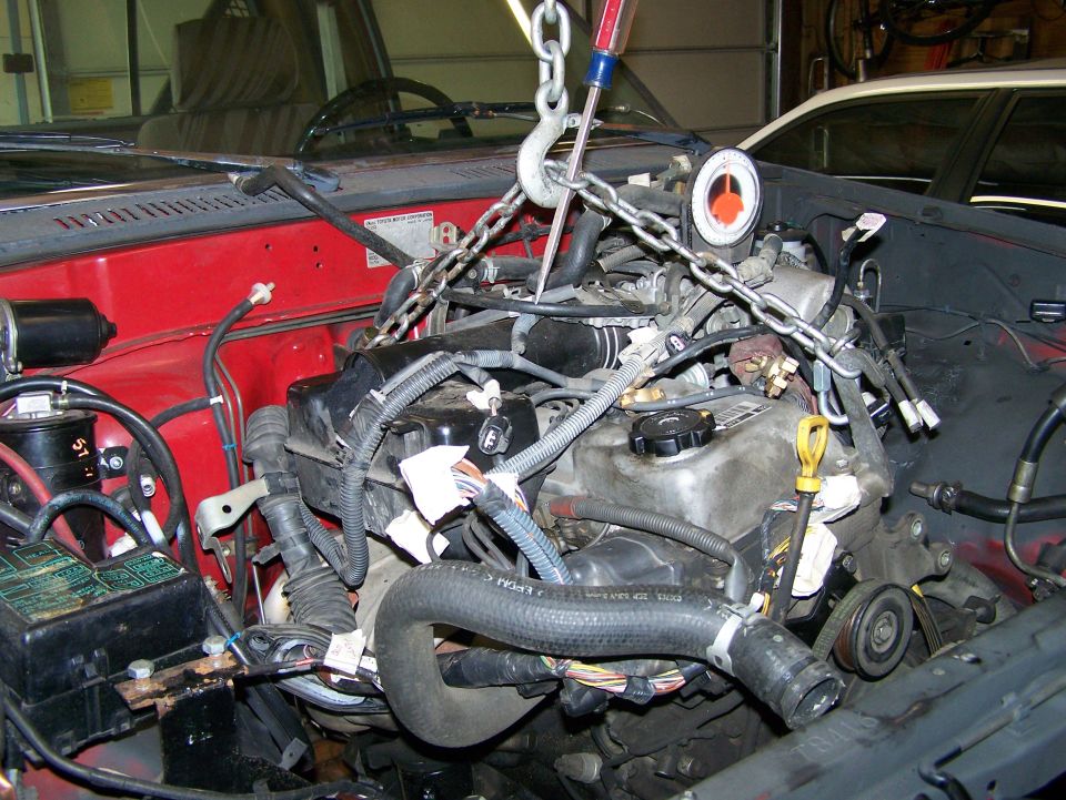 Engine is being lifted out of a red vehicle with chains and a hook. Wires and hoses are connected