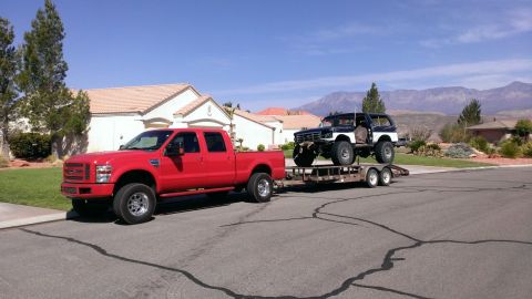 1994 Ford Bronco - A red Ford truck tows a trailer carrying a lifted, dark-colored Bronco with a white top.