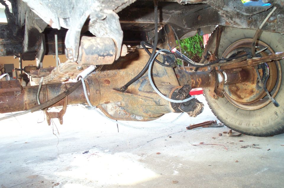 Rusty solid rear axle and driveshaft under a truck, cable-slung for removal