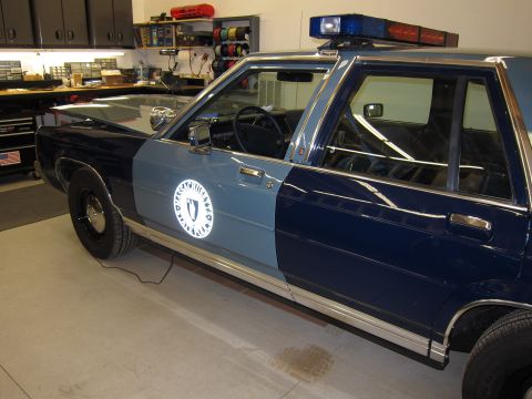 1991 Ford LTD Crown Victoria - A two-tone police car is parked in a garage with shelves of equipment in the background.