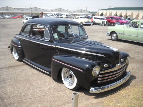 1963 Ford Fairlane - A modified black coupe with a prominent chrome grille and whitewall tires sits in a parking lot with other vehicles.
