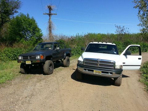 1987 Toyota 4Runner - Two trucks, a lifted dark gray pickup and a white Dodge Ram, are parked on a dirt road under a clear blue sky.