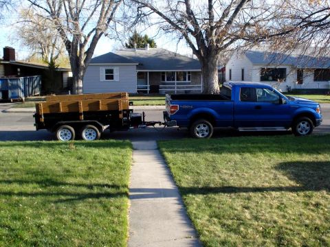 2008 Ford Crown Victoria - A blue pickup truck is parked on the street, hitched to a trailer filled with wood planks.