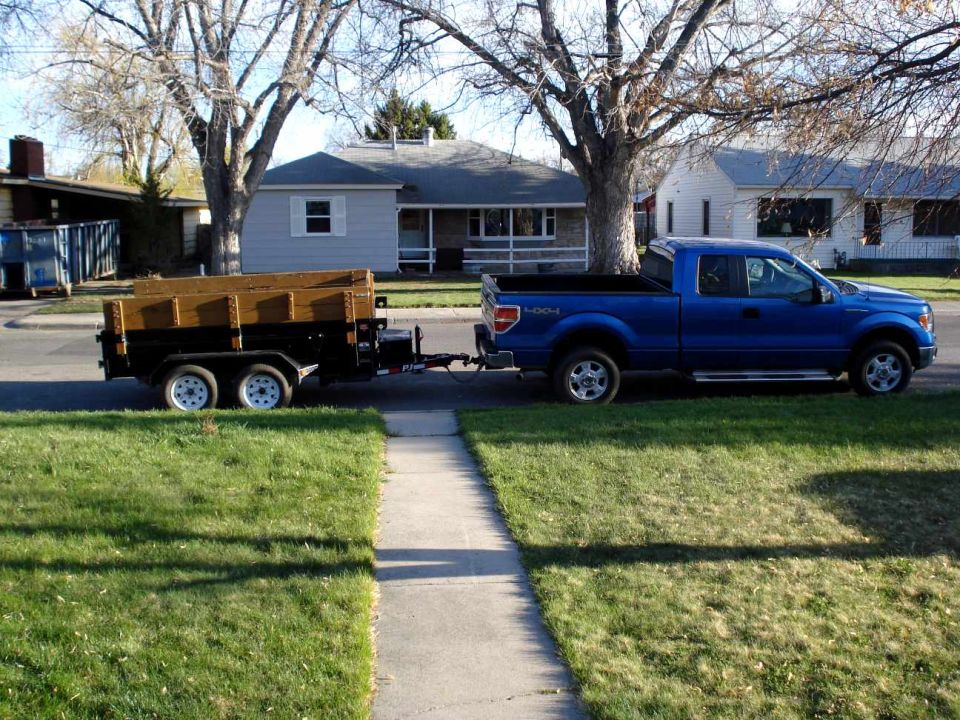 Blue Ford F-150 pickup truck with 4x4 badging, towing a dual-axle utility trailer loaded with lumber