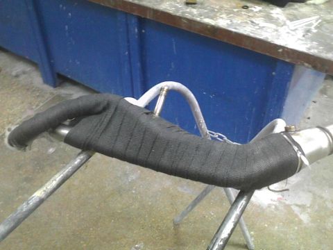 2003 Ford Crown Victoria - A section of exhaust pipe, covered in black heat wrap, is held up by metal stands in a workshop.