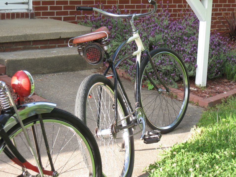 Two bicycles parked on a sidewalk in front of a brick house. One has a red taillight