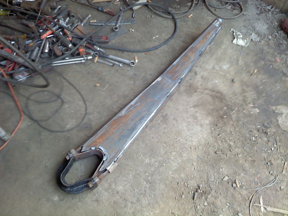 Long metal part with a U-shaped bracket welded to one end, lying on a workshop floor with tools in the background