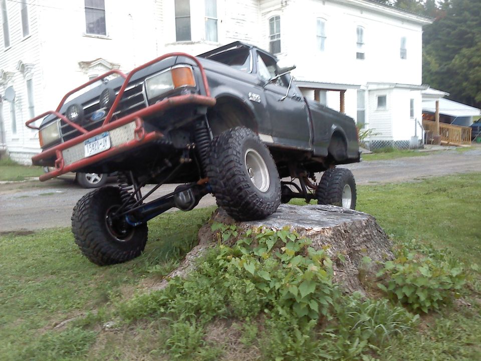 Dark gray Ford F-150 pickup truck with custom red bull bar, lifted suspension, and large tires, parked on a tree stump