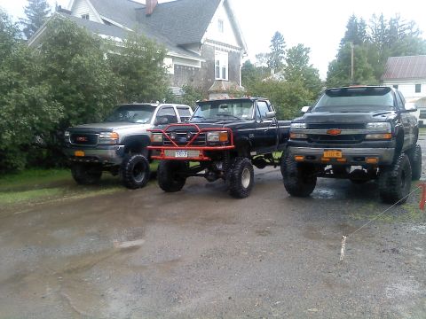 1995 Ford F-150 - Three modified pickup trucks are parked in a gravel lot in front of a house.