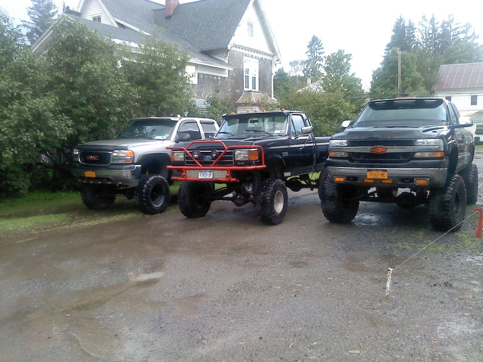 Black Ford F-150 with red bull bar, lifted suspension, and large tires parked next to a GMC and Chevrolet pickup