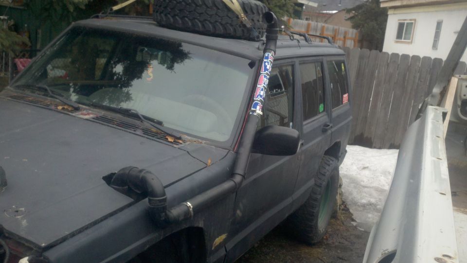 Black Jeep Cherokee with a snorkel kit and spare tire on the roof, parked outside
