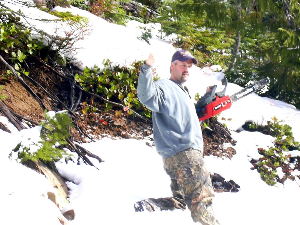 Man in gray sweatshirt and camo pants holding a red chainsaw on a snowy slope