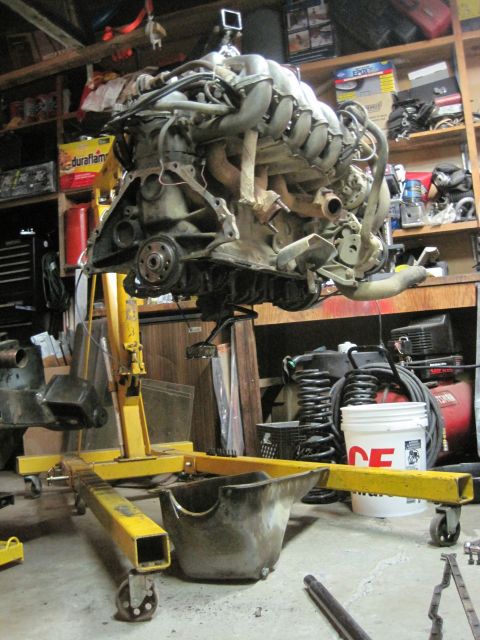 1989 Ford Bronco - An engine is suspended by a yellow engine hoist in a cluttered garage setting.