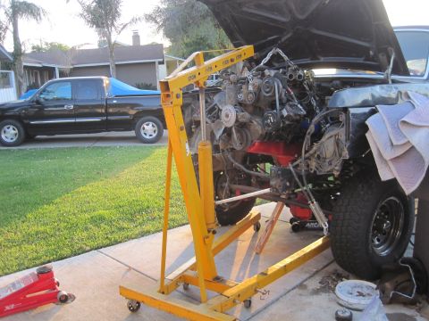 1989 Ford Bronco - Engine hoist is positioned to remove the engine from a vehicle, with a black pickup truck parked nearby.