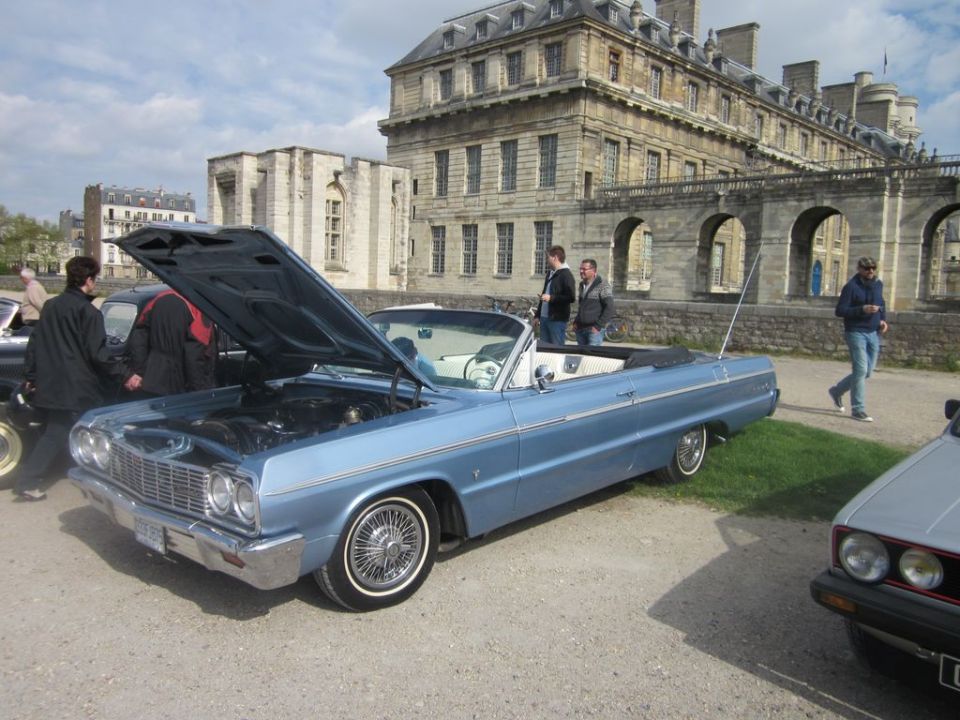 Light blue convertible with hood open, parked in front of a large stone building. Another car is partially visible on the right