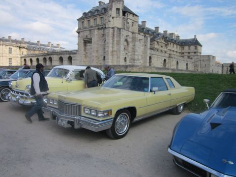 1991 Ford LTD Crown Victoria - A yellow 1975 Cadillac Coupe DeVille is parked in front of a large stone building, with other classic cars nearby.