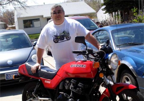 2008 Ford Crown Victoria - A man stands with a red Suzuki motorcycle, a blue car is visible behind him.