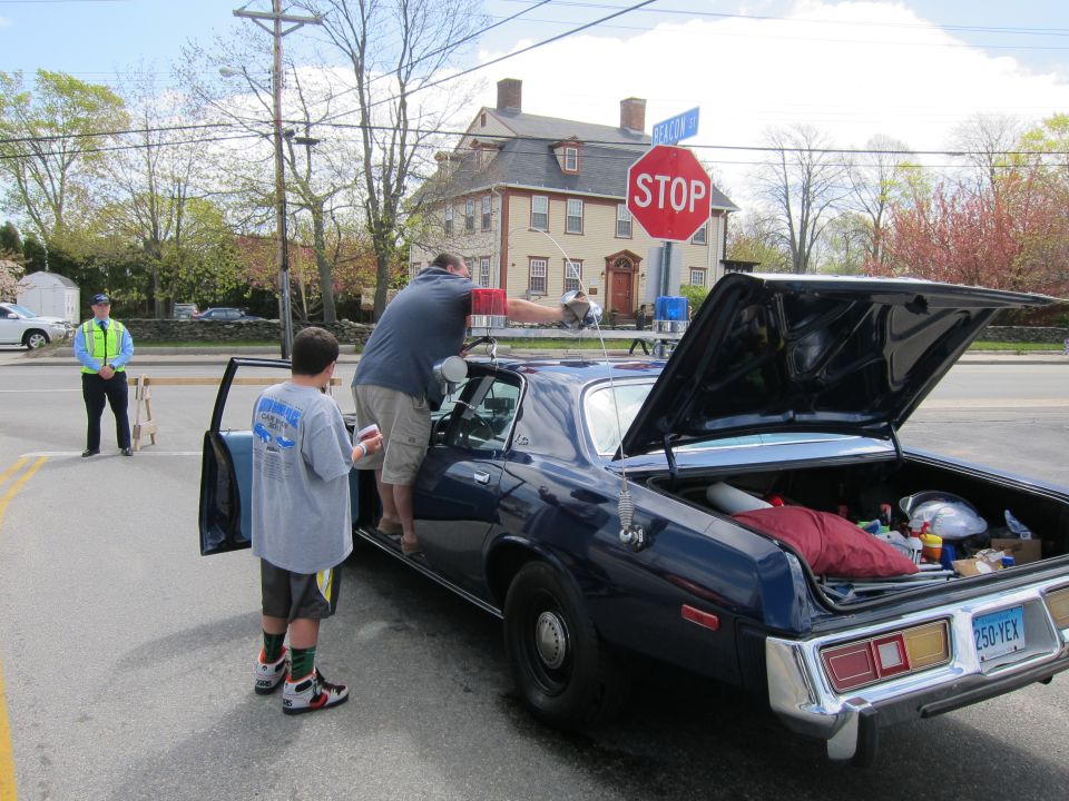 Rear of dark police sedan with open trunk and people, stop sign and brick house behind