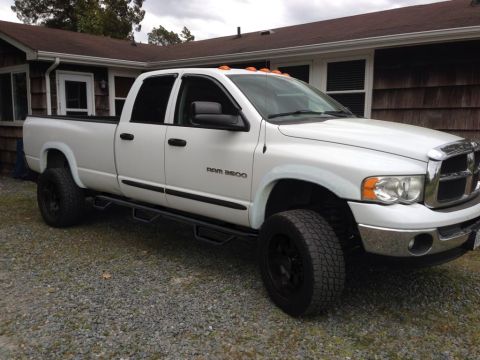 1994 Ford Bronco - White Dodge Ram 3500 quad-cab long-bed pickup on a gravel driveway beside a shingled house, shown from the front three-quarter with RAM 350…