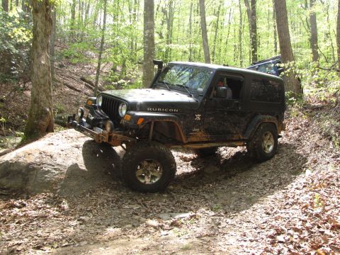 1979 Ford Bronco - A muddy Jeep Wrangler navigates a rocky, wooded trail, with another vehicle following behind.