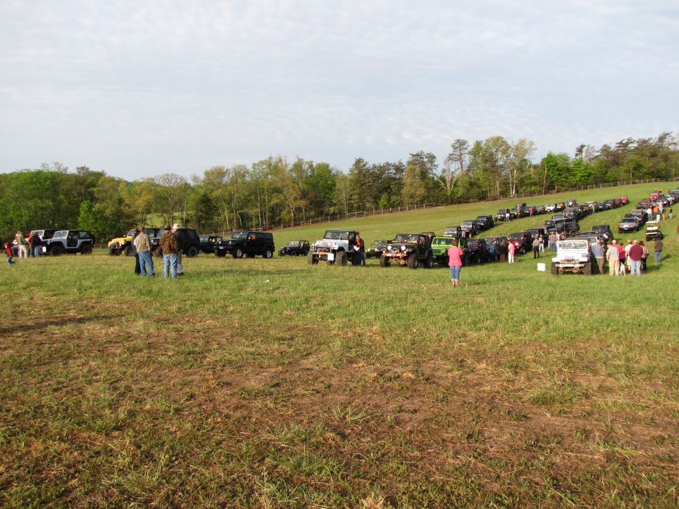 Many off-road vehicles parked in a grassy field with trees in the background